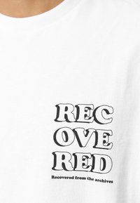 Hvid bomulds T-shirt med fed sort tekst i et legende skrift, der står "RECOVERED" og "Recovered from the archives" med mindre bogstaver.