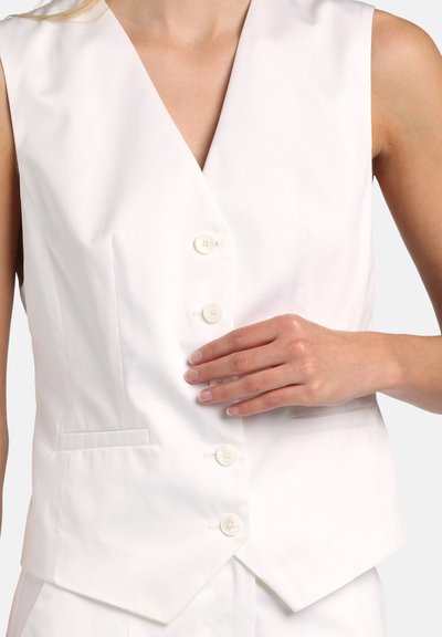 Žena v bielej bezrukávovej veste na gombíky s dvoma prednými vreckami, ruka spočíva na oblasti brucha.