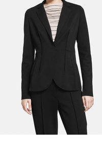 Blazer noir sur mesure avec une silhouette ajustée, tissu en satin, revers crantés et détails de couture subtils. Porté sur un haut rayé.