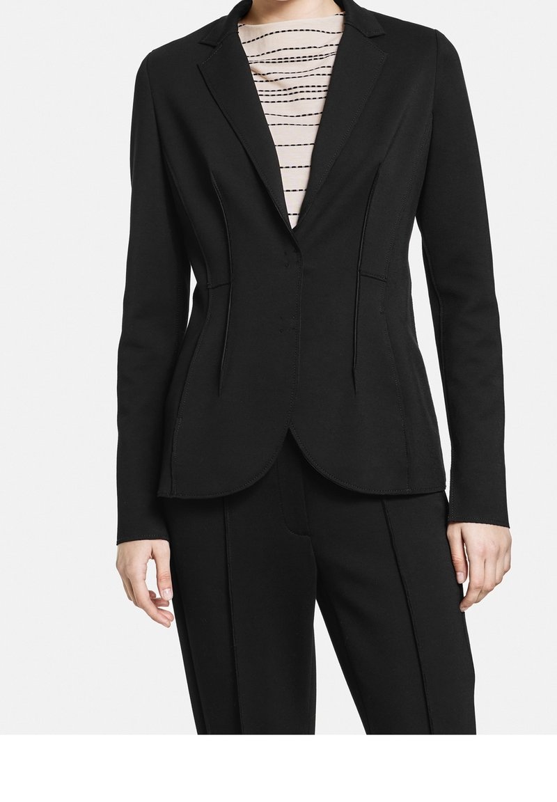 Blazer noir sur mesure avec une silhouette ajustée, tissu en satin, revers crantés et détails de couture subtils. Porté sur un haut rayé.
