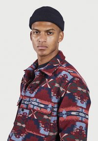 Veste en polaire avec des motifs géométriques rouges, bleus et verts ; dotée d'un col, de deux poches poitrine et de boutons noirs. Portée avec un bonnet bleu marine.