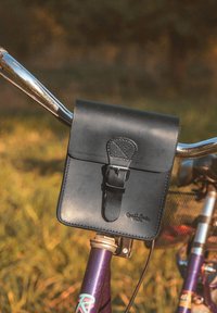 Borsa nera in pelle con chiusura a fibbia attaccata al manubrio di una bicicletta, caratterizzata da una texture liscia e loghi in rilievo sulla parte frontale.