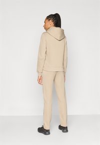 Μπεζ φούτερ με κουκούλα και ταιριαστά παντελόνια φόρμας από απαλό ύφασμα. Διαθέτει κουκούλα με κορδόνι και εφαρμοστά μανίκια. Μαύρα παπούτσια ολοκληρώνουν την εμφάνιση.