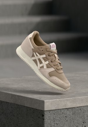 Beige athletic shoe with mesh and suede material, featuring white accents, a cushioned sole, and a pink logo on the tongue.