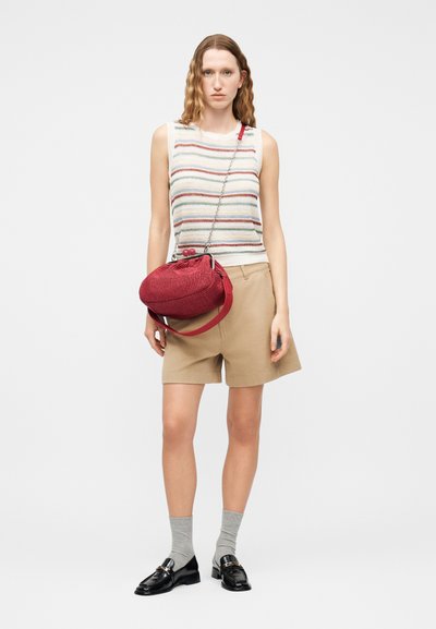 Γυναίκα που φοράει ριγέ πλεκτή μπλούζα χωρίς μανίκια, μπεζ σορτς, γκρι κάλτσες, μαύρα loafers και κρατά μια κόκκινη υφασμάτινη τσάντα χιαστί με αλυσίδα.