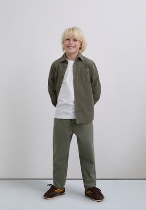 Niño sonriente con cabello rubio que está de pie con las manos detrás de la espalda, usando una camisa blanca, una chaqueta verde oliva de botones, pantalones verdes y zapatillas marrones.