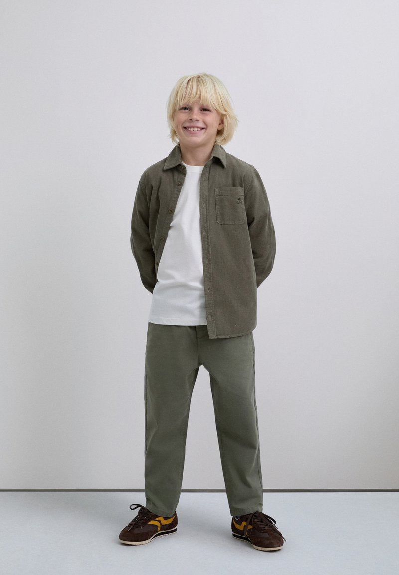 Niño sonriente con cabello rubio que está de pie con las manos detrás de la espalda, usando una camisa blanca, una chaqueta verde oliva de botones, pantalones verdes y zapatillas marrones.