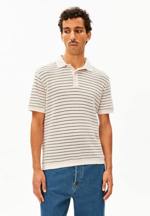 Mann mit lockigem Haar und Schnurrbart, der ein weißes kurzärmeliges gestreiftes Poloshirt und blaue Jeans trägt und vor einem einfarbigen Hintergrund steht.