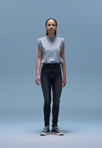 Siva brezrokavnik, črni kavbojke z visokim pasom in črni supergi; na vratu srebrna verižica; nevtralno ozadje.