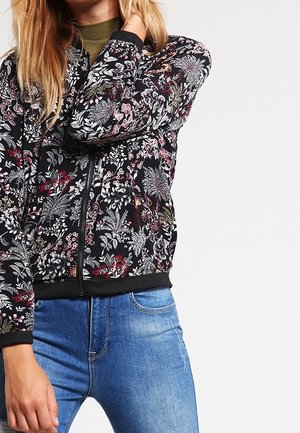 Femme portant une veste zippée noire à fleurs par-dessus un haut vert avec un jean bleu, debout devant un fond blanc uni.