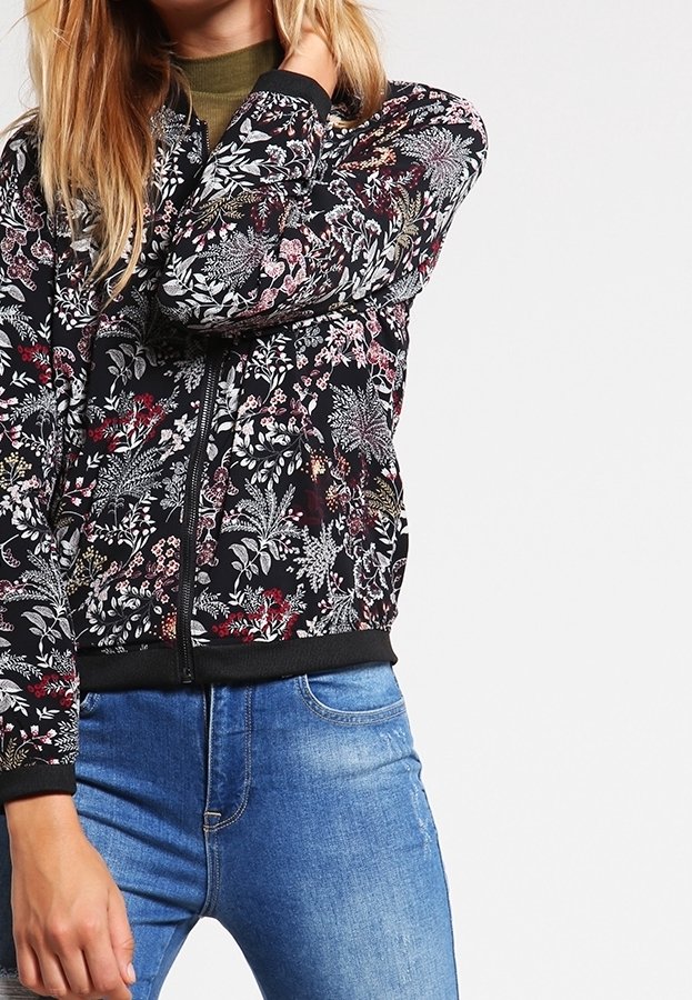 Femme portant une veste zippée noire à fleurs par-dessus un haut vert avec un jean bleu, debout devant un fond blanc uni.