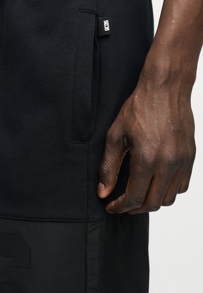 Χέρι με σκούρο δέρμα που ακουμπά κοντά στην πλαϊνή τσέπη μαύρου παντελονιού με μια μικρή μαύρη και λευκή υφασμάτινη ετικέτα με κείμενο.