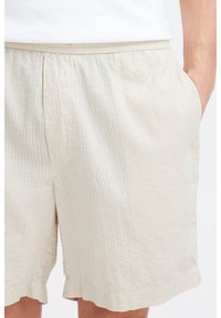 Pantaloni scurți bej deschis dintr-un material texturat, cu talie elastică și buzunare laterale, tivuri ondulate și dungi verticale subtile.