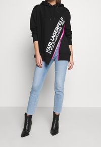 Czarna bluza z kapturem z białym diagonalnym logo i jasnoróżowym detalem zamka błyskawicznego. Połączona z jasnoniebieskimi dżinsami i czarnymi botkami do kostek.