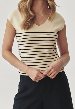 Vrouw draagt een mouwloze crèmekleurige top met zwarte horizontale strepen en een donkere broek, handen gevouwen voor zich.