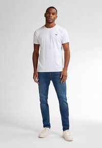 Biała koszulka z krótkim rękawem z małym logo na lewej piersi, zestawiona z niebieskimi dżinsami i jasnymi trampkami. Minimalistyczny design.