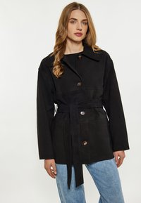 Cappotto nero con cintura realizzato in tessuto morbido, dotato di un colletto, bottoni frontali e una vestibilità comoda, abbinato a jeans blu chiaro.