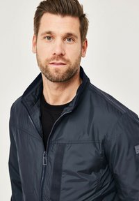 Navy blue zip-front jacket with high collar, featuring a smooth texture, horizontal seam details, and a small logo on the left shoulder.