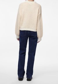 Krämfärgad stickad tröja med texturerat mönster, vida ärmar och ribbad nederkant, i kombination med blå denimjeans och svarta skor.