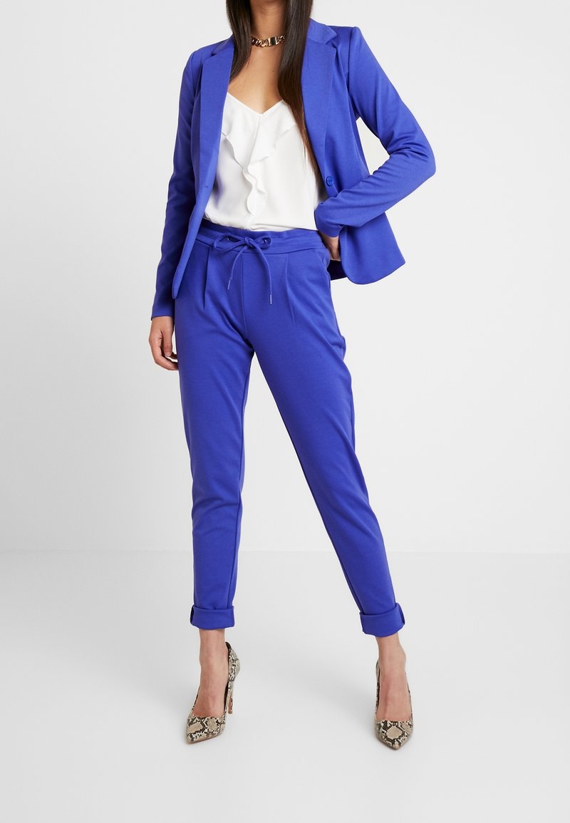 Femme portant un blazer bleu et un pantalon à cordon assorti avec des revers roulés, un chemisier blanc à volants, des escarpins imprimé serpent et un collier en or.