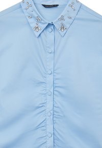 Camicia button-up azzurro chiaro con davanti arricciato, caratterizzata da un colletto decorativo adornato con gemme trasparenti. Tessuto morbido e leggero.