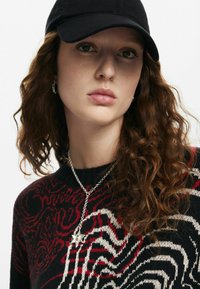 Cappello nero, capelli castani ricci, primo piano di una persona che indossa un maglione a fantasia nero e rosso, collana d'argento con un pendente a forma di stella.