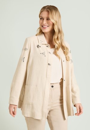 Blonde Frau lächelnd, trägt eine cremefarbene Jacke mit silbernen Blumenverzierungen, passende Hose und ein helles Oberteil vor einem schlichten Hintergrund.