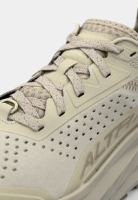 Baskets de course beiges claires avec une tige en maille, un design perforé, des lacets contrastés et une semelle texturée. Présente un logo sur le côté.