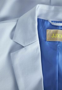 Helles blaues Blazer mit glatter Textur, ausgestattet mit einem strukturierten Kragen und einem kontrastierenden blauen Innenfutter. Etikett innen sichtbar.