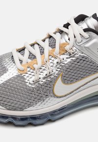 Zapatilla plateada y gris con parte superior de malla, cordones blancos y detalles dorados. Cuenta con una suela visible con amortiguación de aire y el logotipo de Nike.