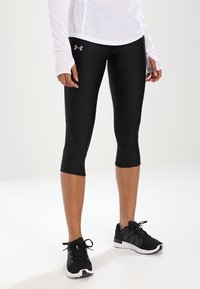 Czarne legginsy capri wykonane elastycznego materiału, o dopasowanym kroju z detalami szwów. W zestawieniu z czarnymi butami sportowymi.