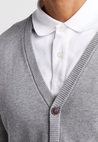 Cardigan grigio con design a V, caratterizzato da bordi a coste e un singolo bottone marrone. Indossato sopra una camicia bianca con colletto.
