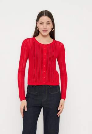 Frau trägt eine enganliegende rote, geknöpfte Strickjacke mit vertikalen Lochmustern und dunkle hoch taillierte Jeans, steht vor einem weißen Hintergrund.