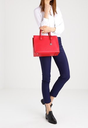 Červená kožená kabelka s texturovaným povrchem, dvěma horními uchy a kovovým zámkem. Kombinována s tmavě modrými kalhotami a černými botami.