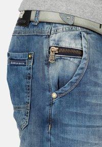 Jean en denim bleu avec une finition décolorée, doté d'une poche zippée, d'accents de couture jaunes et d'un bouton en argent sur la poche avant.