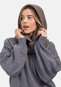 Kobieta w szarej, oversize'owej bluzie i grubym dzianinowym kapturze w kolorze taupe, poprawiająca kaptur i patrząca w bok.