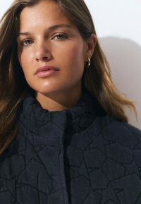 Veste matelassée bleu foncé avec col haut, présentant un motif géométrique et un léger éclat. Le modèle porte des boucles d'oreilles en forme de créoles dorées.