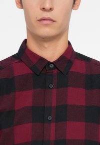 Primer plano de una persona con una camisa de cuadros roja y negra, con el cuello abrochado, y una expresión facial neutral visible desde la nariz hacia abajo.