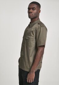 Camisa de manga corta color verde oliva, fabricada en una tela suave, que cuenta con dos bolsillos en el pecho, botones y hombreras.