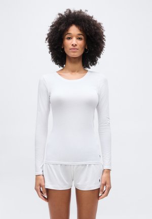 Woman with curly hair wearing a plain white long-sleeve shirt and white shorts, standing against a plain white background.