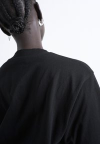 Personne à la peau foncée et aux cheveux tressés attachés en chignon, portant des boucles d'oreilles argentées en forme d'anneau et une chemise noire, vue de dos sur un fond blanc.