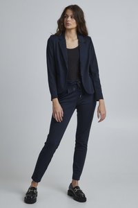 Marineblazer mit einem einzigen Knopf, kombiniert mit marineblauen Jogginghosen. Schwarzes Oberteil darunter, getragen mit schwarzen Plateauslippern.