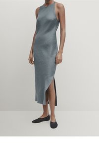 Robe sans manches, longueur cheville, en tissu bleu ardoise texturé, avec une fente sur le côté et une silhouette ajustée. Associe avec des chaussures plates noires.