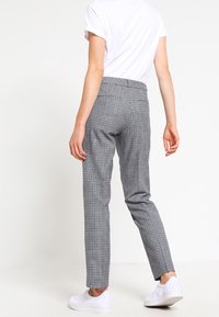 Person som bär grå rutiga byxor, vit t-shirt och vita sneakers, står med ryggen mot kameran på vit bakgrund.