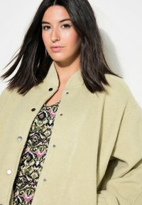 Lichtgroene, oversized fleece jas met een hoge kraag en drukknopen, gelaagd over een patroon top met roze en zwarte accenten.