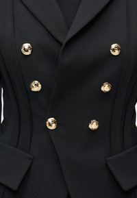 Μαύρο διπλόμπροστο μπλέιζερ με έξι χρυσά κουμπιά, δομημένες ραφές και τσέπες με καπάκι, φορεμένο πάνω από μαύρο τοπ.