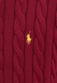 Röd kabelstickad tröja gjord av strukturerat stickat tyg, med en liten gul polo-spelare logotyp som accent på framsidan.