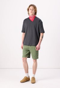 Pelēks īsām piedurknēm T-krekls ar brīvu piegriezumu, zaļas šorti un sarkana bandana. Modelis valkā brūnus apavus un baltas zeķes.