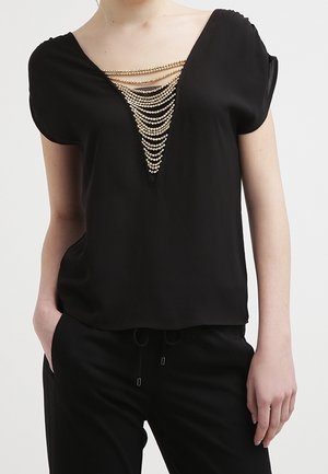 Kvinde iført sort kortærmet bluse med dyb V-hals udsmykket med kaskader af guldkæde-detaljer, kombineret med sorte bukser.