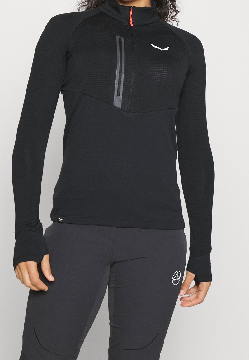 Svart långärmad topp med kvartzip, texturerad övre panel, tumhål och en ficka med dragkedja på bröstet. Har en logotyp på bröstet.